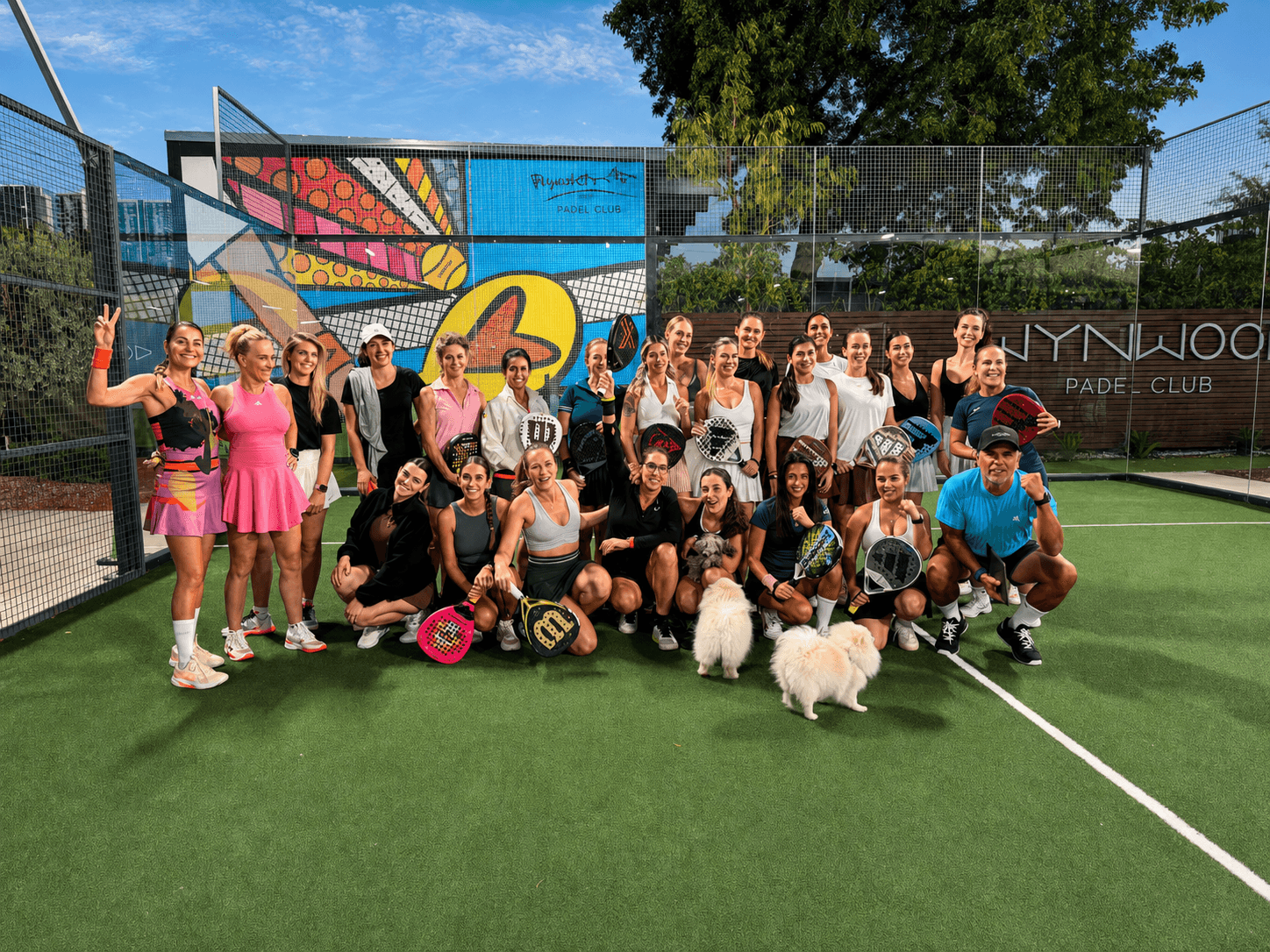 A group of women members posing on court in front of the Wynwood mural wall with two small dogs