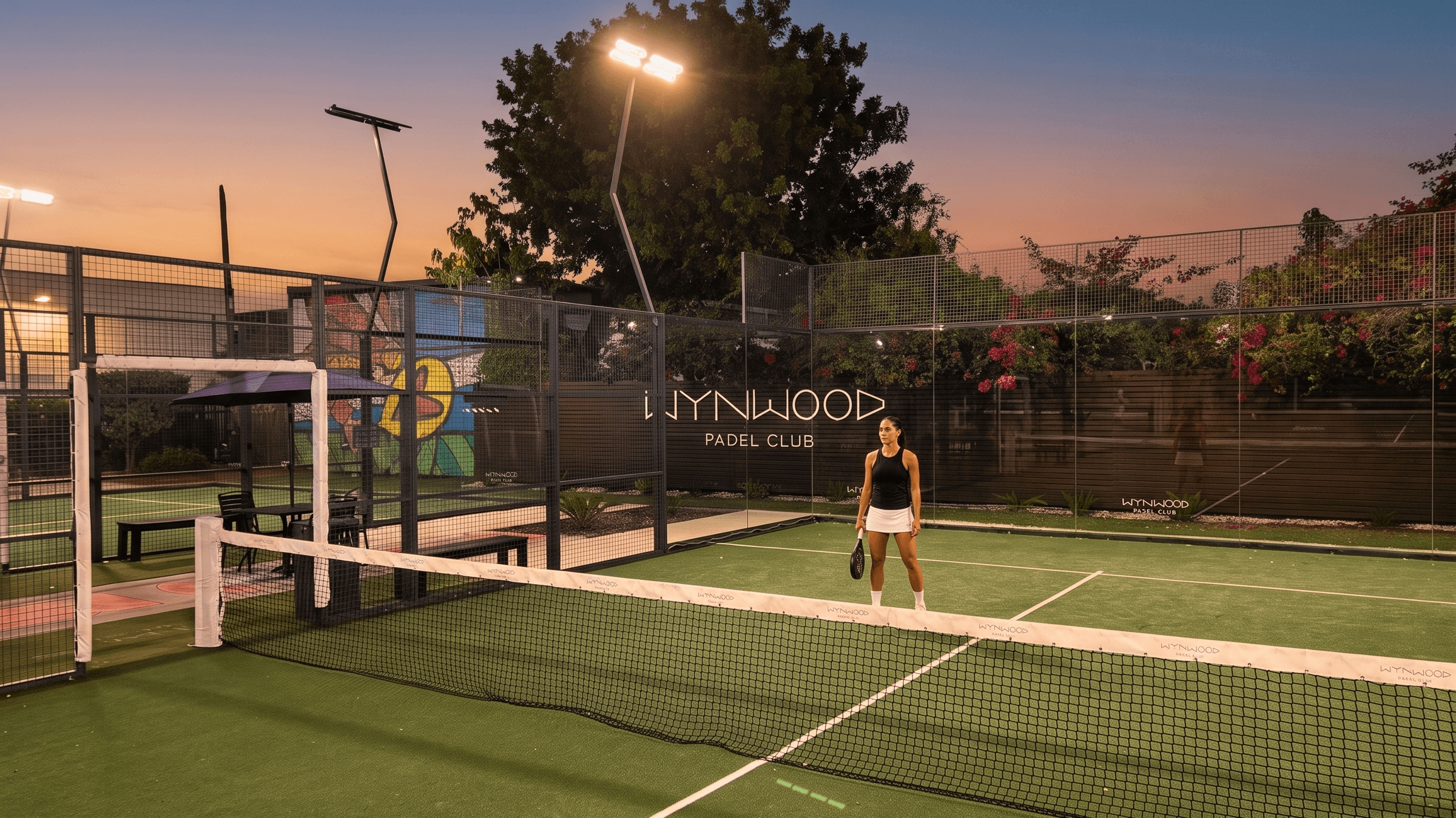 A player standing on the padel court at dusk, Wynwood Padel Club signage lit up behind her